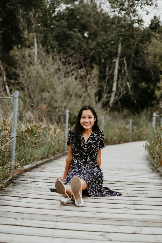 a woman sitting on top of a wooden walkway, a picture, inspired by helen huang, unsplash, happening, happily smiling at the camera, wearing a dress, low quality photo, smol