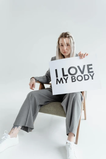 a woman sitting in a chair holding a sign that says i love my body, sydney sweeney, profile image, profile picture, humanoid body