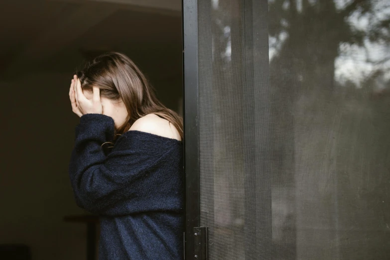 a woman standing in front of a window covering her face, pexels, in a navy blue sweater, devastated, leaning on door, brunette woman