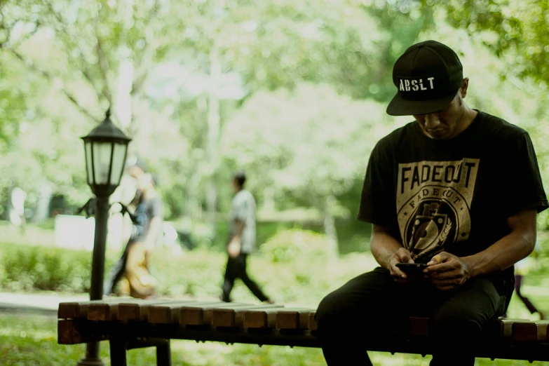 a man sitting on a bench looking at his cell phone, inspired by Rudy Siswanto, realism, outlive streetwear collection, faded hat, sullen, in a park