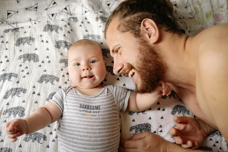 a man laying on top of a bed next to a baby, pexels contest winner, happening, beard stubble, cheeky smile, patterned, thumbnail