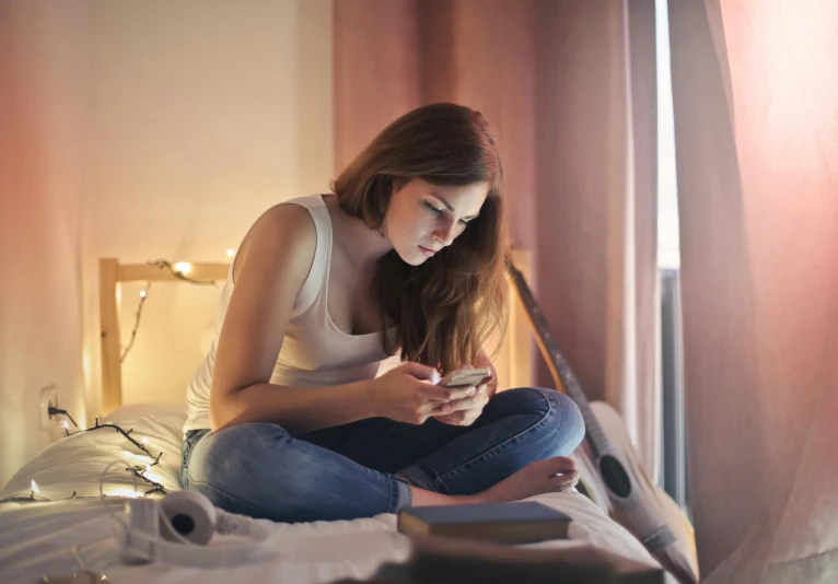 a woman sitting on a bed looking at her phone, trending on pexels, lights on, student, sitting on the floor, low