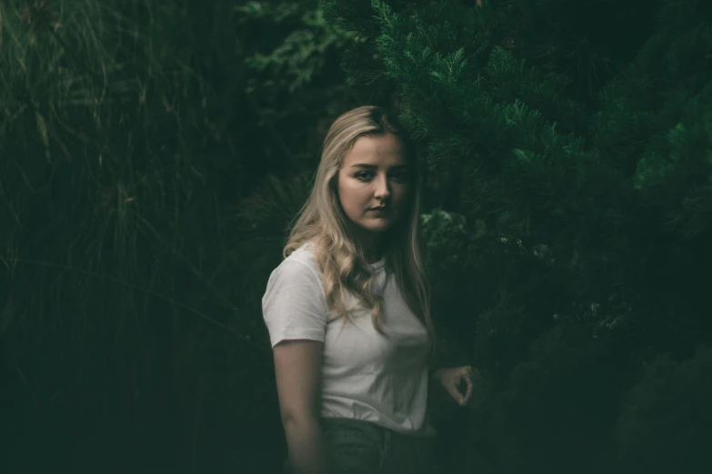 a woman standing in the middle of a forest, inspired by Elsa Bleda, unsplash contest winner, realism, portrait of kim petras, dressed in a white t shirt, dark green tones, ((portrait))