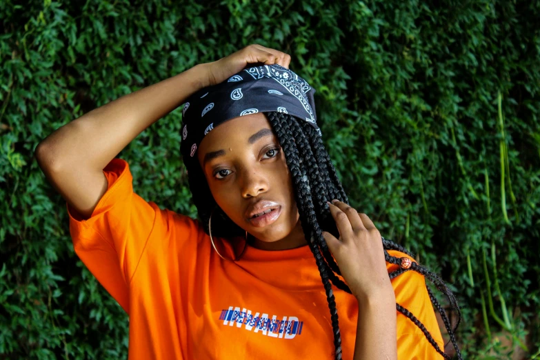 a close up of a person wearing an orange shirt, an album cover, by Bernardino Mei, trending on pexels, visual art, black hair in braids, bandana, black teenage girl, outlive streetwear collection