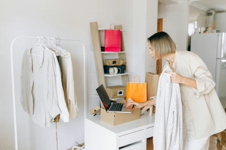 a woman standing in front of a desk with a laptop, by Nicolette Macnamara, trending on pexels, happening, exiting from a wardrobe, cardboard, shopping groceries, white and gold robes