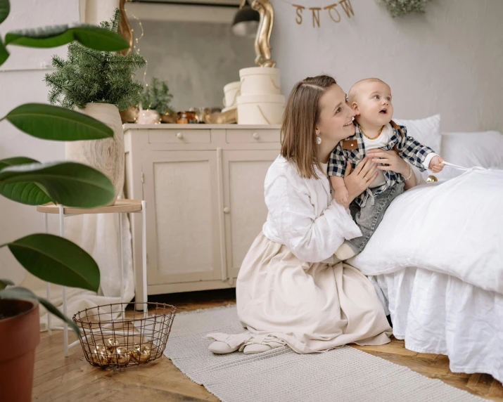 a woman sitting on a bed holding a baby, pexels contest winner, decoration around the room, white, 1 4 9 3, small