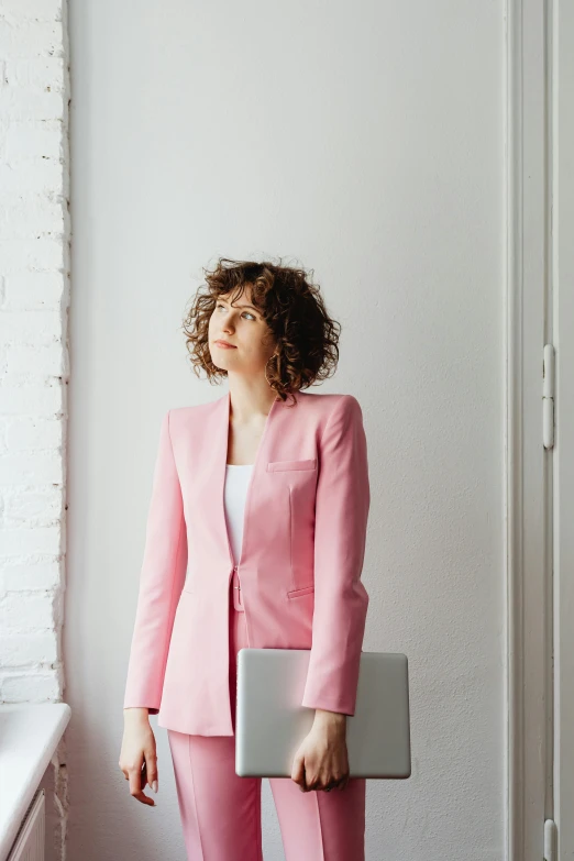 a woman standing in front of a window holding a laptop, an album cover, by Miroslava Sviridova, trending on pexels, renaissance, wearing a pink tux, curly haired, ready for a meeting, business attire