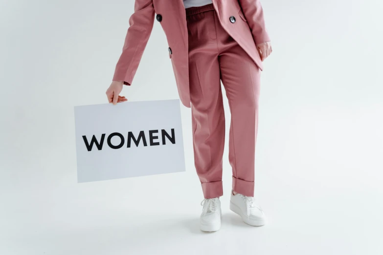 a woman in a pink suit holding a sign, trending on pexels, womanhood, wearing pants, trench coat and suit, white woman