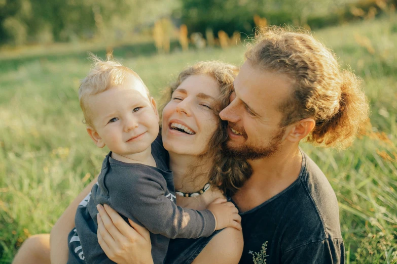 a man sitting next to a woman holding a baby, by Adam Marczyński, pexels contest winner, hugging each other, avatar image, small smile, beautiful daylight