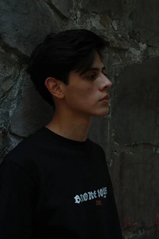 a man standing in front of a stone wall, an album cover, pexels contest winner, he is wearing a black t-shirt, nonbinary model, profile image, portrait of 1 5 - year - old boy