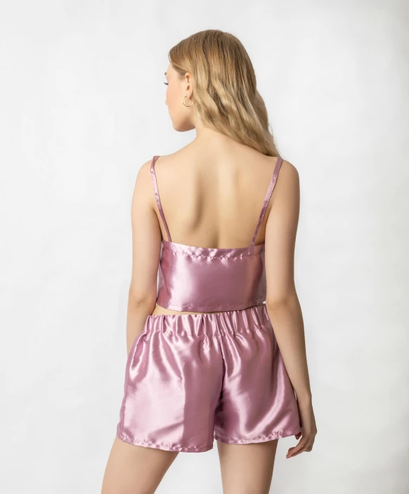 a woman wearing a pink satin romper, by Nina Hamnett, bralette, sitting on the edge of a bed, 3 / 4 back view, square
