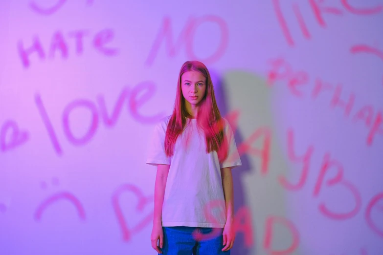 a girl standing in front of a wall with writing on it, pexels, love hate love, pink violet light, portrait of white teenage girl, male teenager