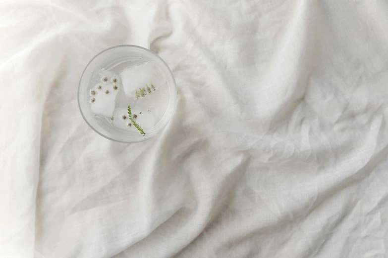 a glass of water sitting on top of a bed, inspired by Vija Celmins, trending on pexels, renaissance, white splendid fabric, botanicals, light scatter, detailed product image