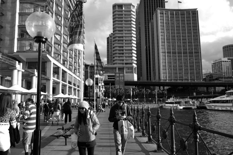 a group of people walking down a sidewalk next to a river, a black and white photo, inspired by Thomas Struth, hurufiyya, sydney, big buildings, vivid!!, harbour