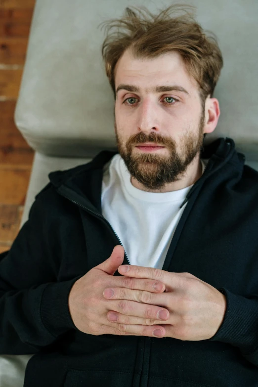 a man with a beard sitting on a couch, lysergic, looking down on the camera, resting on chest, hands straight down