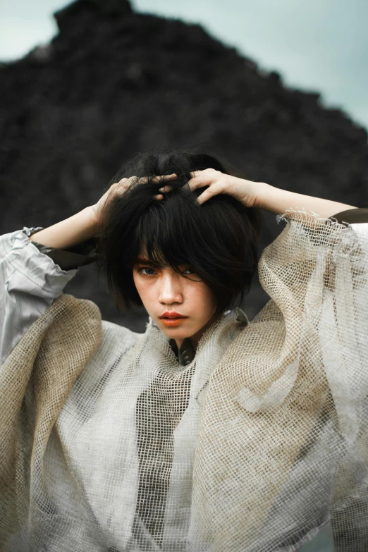 a woman standing in front of a mountain with her hands on her head, an album cover, inspired by Itō Shinsui, unsplash, messy black bob hair, wearing desert poncho, beautiful grumpy girl, a photo of a disheveled man