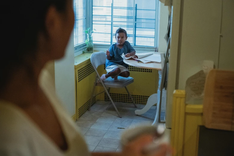 a woman sitting in a chair in front of a window, by Lily Delissa Joseph, pexels contest winner, with a kid, seated at a table, documentary still, joel torres