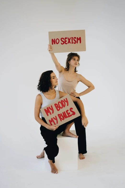 two women holding a sign that says no sexism, an album cover, trending on pexels, feminist art, body portrait, hila klein, studio photo, actors