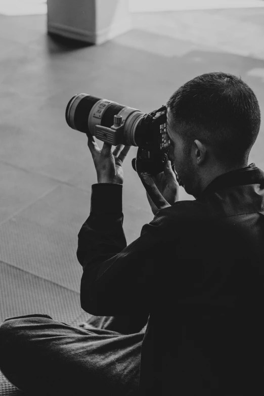 a man sitting on the floor taking a picture with a camera, a black and white photo, by Adam Marczyński, telephoto zoom, looking from shoulder, professional photo-n 3, 4k. profession photography