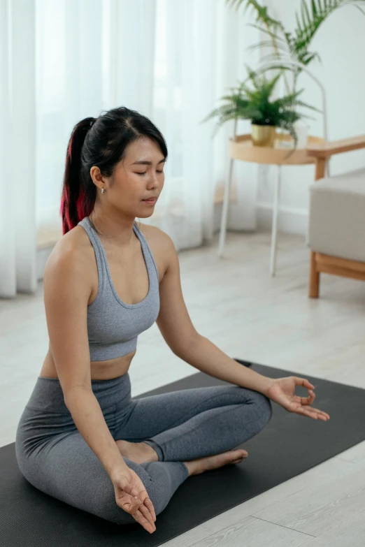 a woman sitting on a yoga mat in a living room, wearing a light grey crown, asian features, hard breathing, darren quach