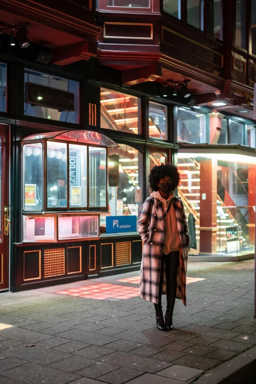 a woman walking down a sidewalk at night, shop front, red building, afro - futurist style, cosy vibes