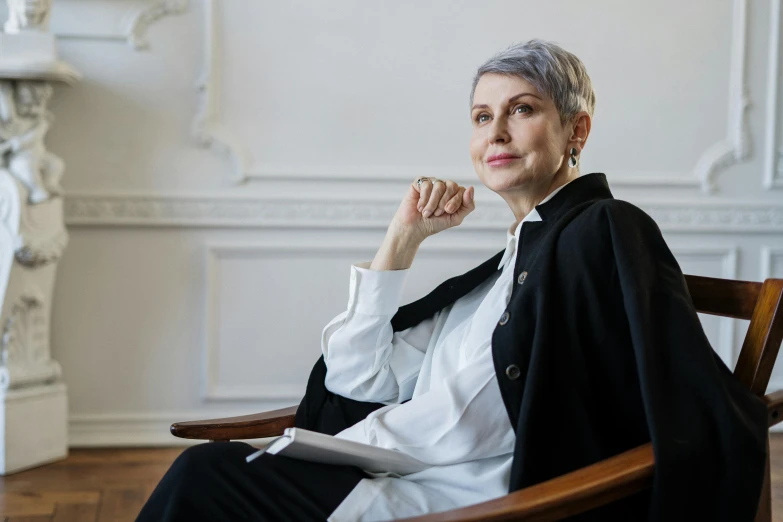 a woman sitting in a chair in a room, a portrait, pexels contest winner, photorealism, short silver hair, wearing an academic gown, jovana rikalo, androgynous person