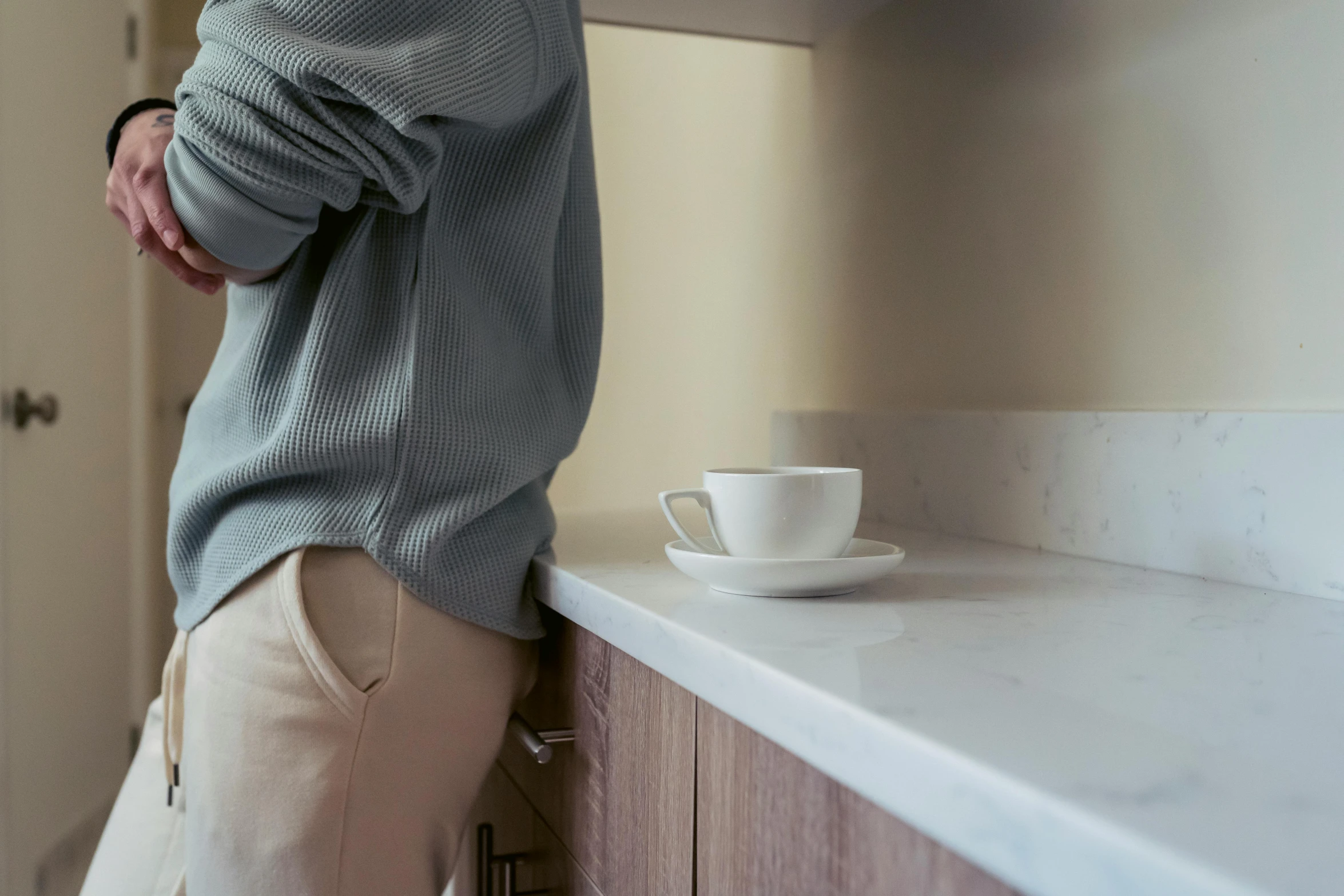 a man standing in a kitchen with a cup of coffee, happening, wearing pants, grey, hands on counter, subtle detailing