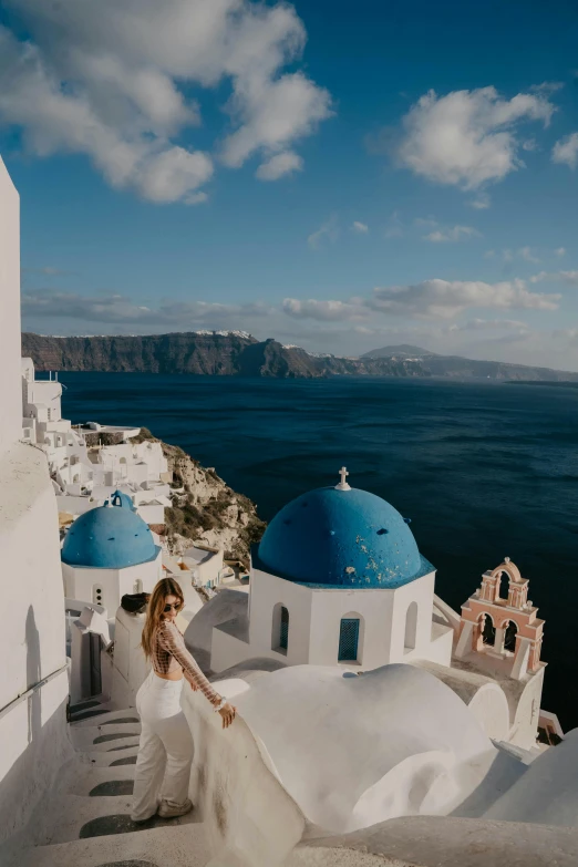 a woman standing on top of a white building next to the ocean, by Alexis Grimou, trending on unsplash, renaissance, with great domes and arches, blue accents, greek fabric, volcano in the background