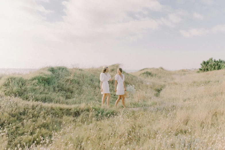 a couple of women standing on top of a grass covered field, unsplash, girl walking between dunes, medium format. soft light, coastal, white