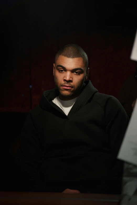 a man sitting at a table in a dark room, an album cover, by Stokely Webster, realism, angry looking at camera, photograph credit: ap, in a black hoodie, jordan peele's face