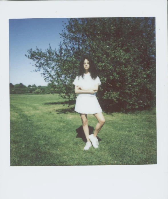 a woman standing on top of a lush green field, a polaroid photo, very pale skin, next to a tree, wearing white clothes, outside in parking lot