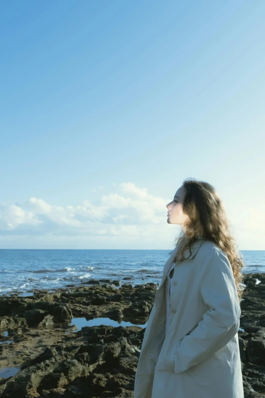 a woman standing on a rocky beach next to the ocean, an album cover, inspired by Wilhelm Hammershøi, unsplash, happening, blue sky, distant thoughtful look, backlit shot girl in parka, profile image