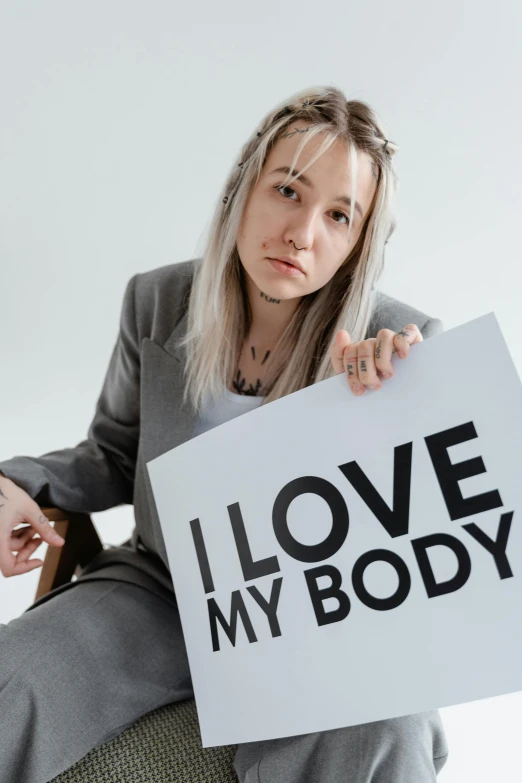 a woman sitting on a chair holding a sign that says i love my body, pexels contest winner, antipodeans, charlie immer and jenny saville, profile image, cruelty, saoirse ronan