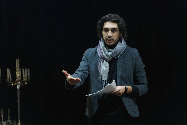 a man in a suit and scarf holding a piece of paper, by Maryam Hashemi, unsplash, renaissance, performing on stage, adam driver, teaching