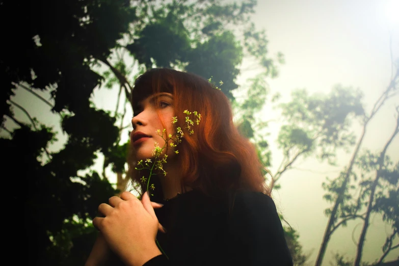 a woman holding a flower in front of her face, an album cover, inspired by Elsa Bleda, magical realism, ( redhead, with a yellow green smog sky, photo realistic”