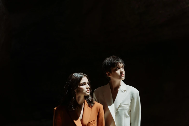 a man and a woman standing next to each other, an album cover, by Emma Andijewska, young women, dark sienna and white, in a cave. underexposed, brunettes