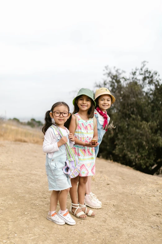 three little girls standing next to each other on a dirt road, unsplash, wearing sunglasses and a cap, los angeles ca, young asian girl, colorful”