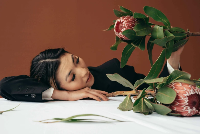 a woman laying on top of a table next to a flower, inspired by Fei Danxu, gemma chan, press shot, next to a plant, looking off into the distance