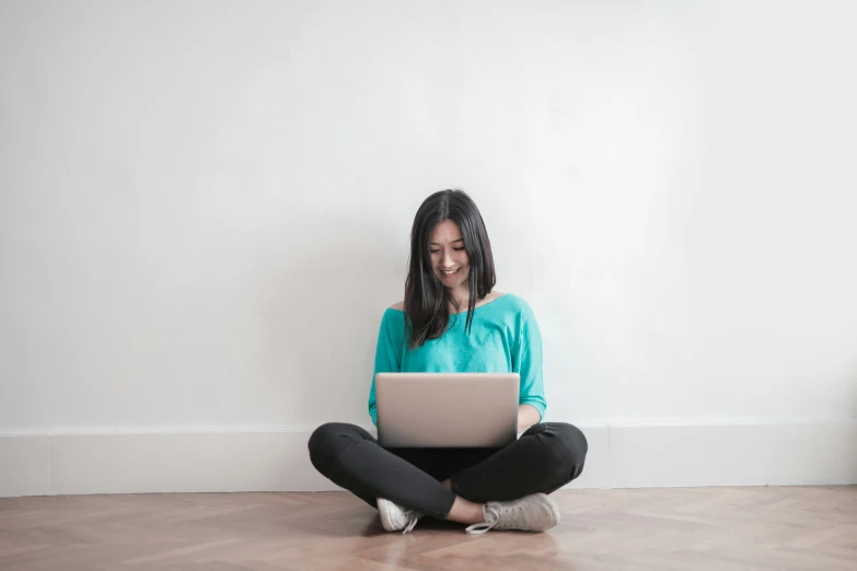 a woman sitting on the floor using a laptop, pexels contest winner, with teal clothes, in white room, subject is smiling, a young asian woman