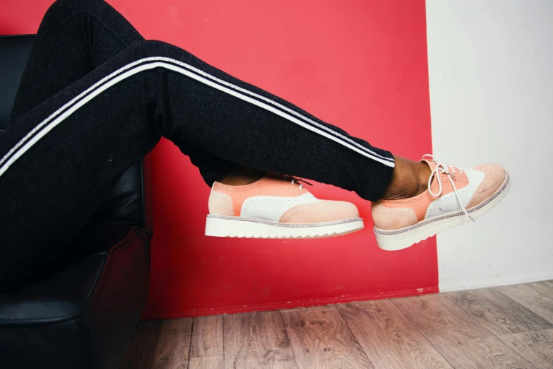 a woman sitting on top of a black chair next to a red wall, trending on pexels, figuration libre, sneaker shoes, toned orange and pastel pink, trimmed with a white stripe, two buddies sitting in a room