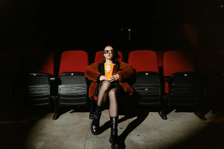 a woman sitting in front of a row of red chairs, a portrait, inspired by Elsa Bleda, pexels contest winner, black and orange coat, drive in movie theater, charli xcx, [ theatrical ]