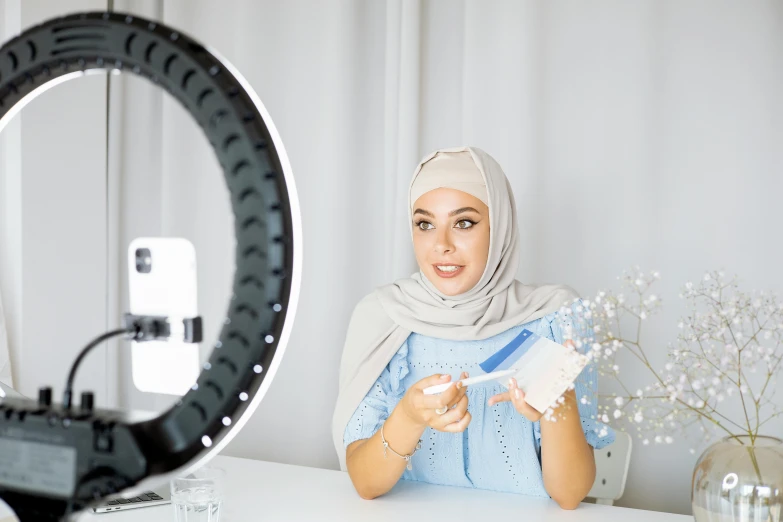 a woman sitting at a table in front of a camera, inspired by Maryam Hashemi, pexels contest winner, hurufiyya, ring light, with a white background, taking a selfie, cardboard