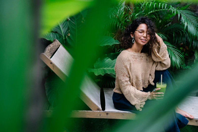 a woman sitting on a bench holding a drink, pexels contest winner, plants and jungle, curly haired, avatar image, full frame image