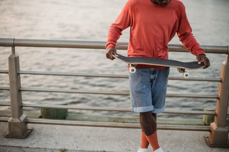 a man standing next to a railing holding a skateboard, pexels contest winner, man is with black skin, at the waterside, wearing an orange t shirt, wearing kneesocks