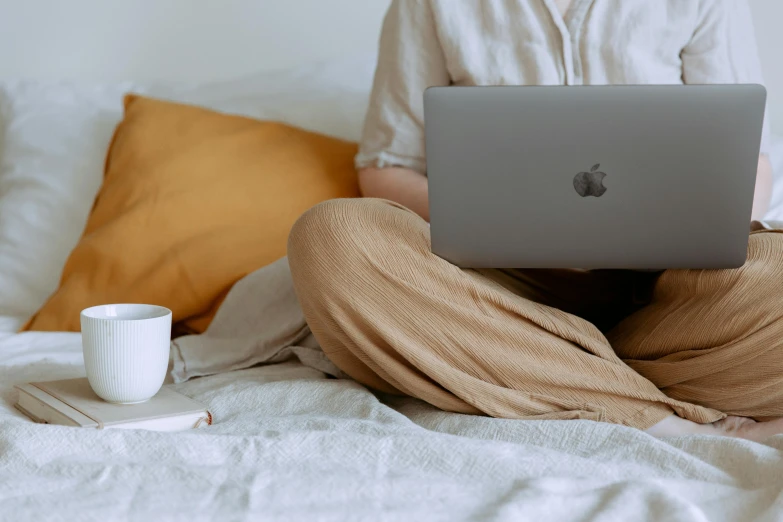 a person sitting on a bed with a laptop, by Carey Morris, trending on pexels, beige, table in front with a cup, from waist up, coloured