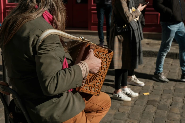 a woman sitting on a bench playing a musical instrument, an album cover, by Raphaël Collin, pexels contest winner, holding a leather purse, intricate carved wood, people walking down a street, close-up photo