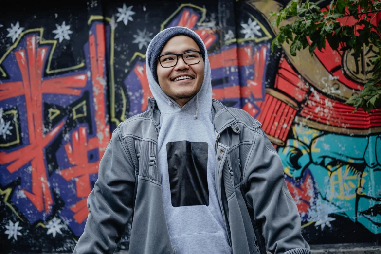 a man standing in front of a graffiti covered wall, a portrait, inspired by Gang Hui-an, unsplash, graffiti, wearing a grey hooded sweatshirt, friendly smile, background image, manuka
