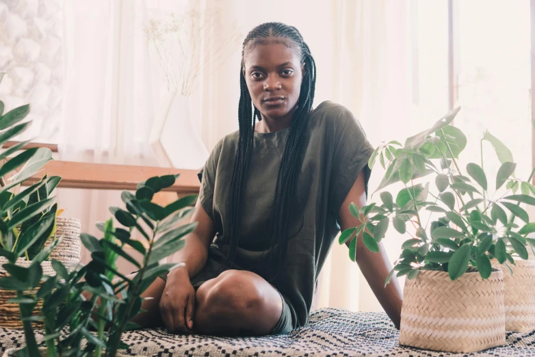 a woman sitting on a bed next to a potted plant, by Dulah Marie Evans, hurufiyya, in a dark green polo shirt, long black braids, nuri iyem, natural lights