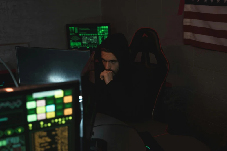 a man sitting at a desk in front of a computer, wearing a dark hood, cryptidcore, immersed within a glitch network, looking distracted