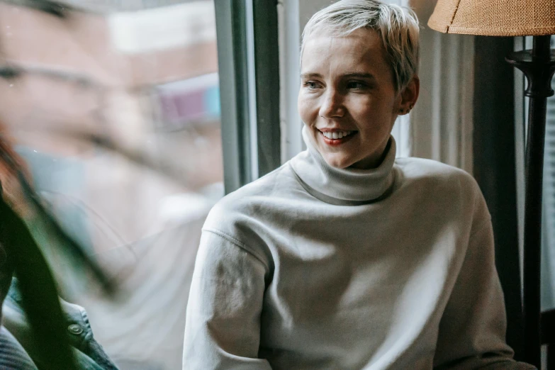 a woman sitting in front of a window smiling, inspired by Nína Tryggvadóttir, pexels contest winner, purism, white short hair, wearing turtleneck, pale grey skin, casually dressed